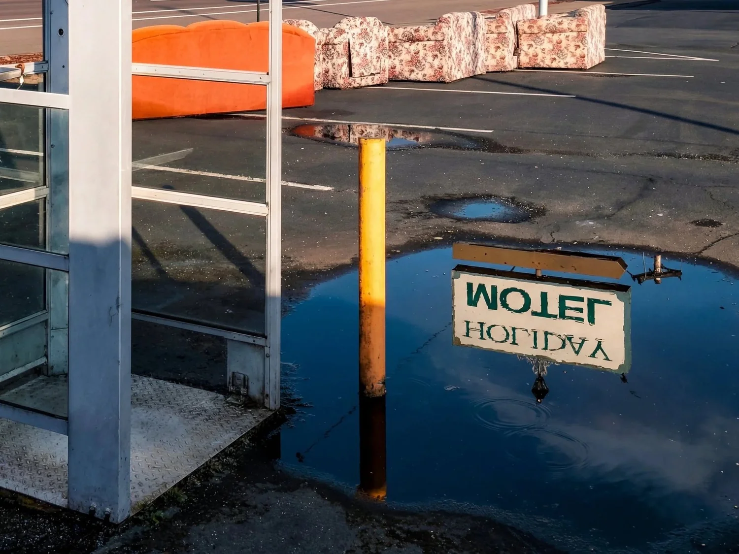 The image shows a parking lot with rain puddles, an orange barrier, floral-patterned couches placed in the lot, a yellow pole, and a mirrored sign that says 'VACANT' in reverse.