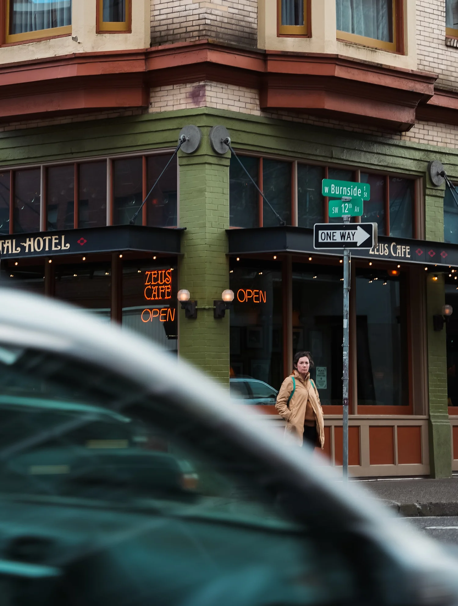 A woman standing outside a corner building with a sign that reads 'Zeus Cafe' and 'Open' in neon lights. Street signs indicate the intersection of W Burnside and SW 12th Avenue, with a one-way street sign. The building has green and beige brick walls