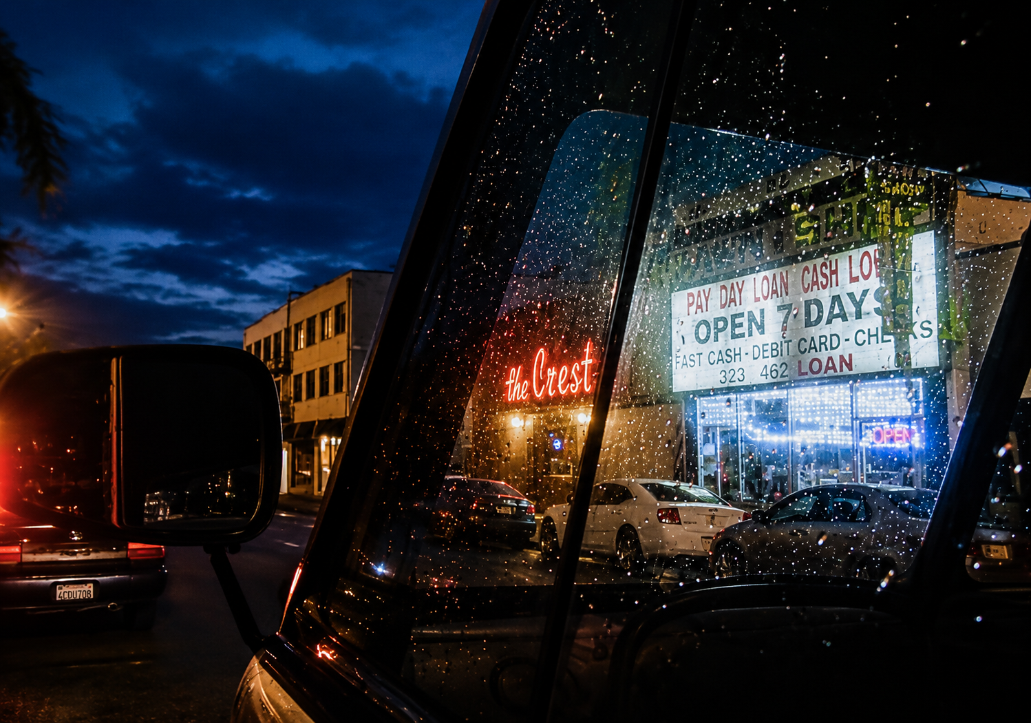 Nighttime scene through rain-streaked car window reflecting neon signs advertising a loan store and a bar, with parked cars and a building on the street