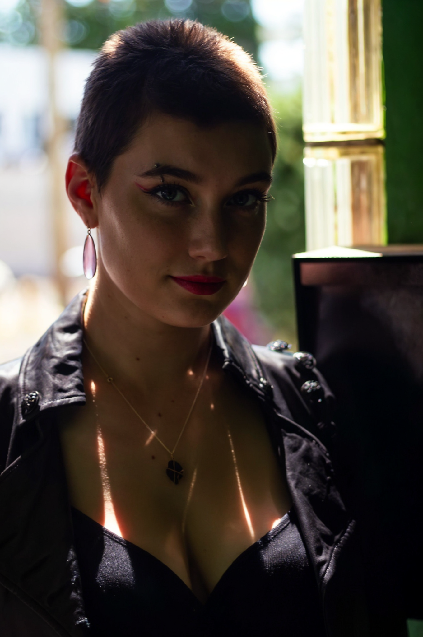 A woman with short dark hair, blue eyes, red lipstick, and eye makeup, wearing large earrings and layered necklaces, standing indoors near an open window with sunlight highlighting part of her face and chest.
