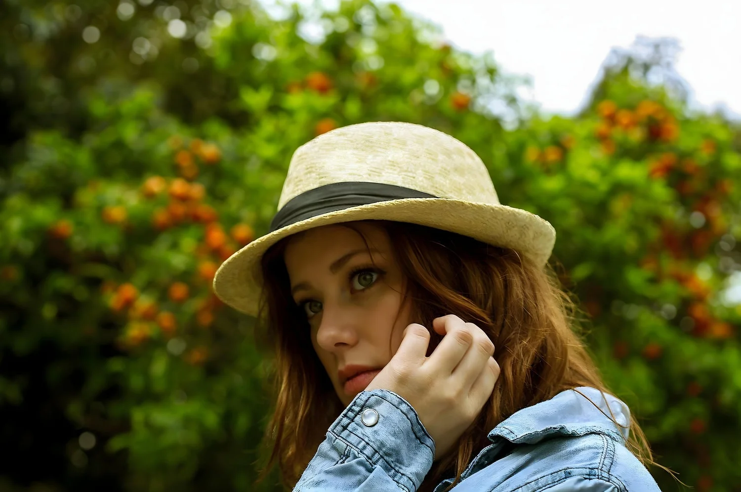 A woman with red hair wears a beige straw hat with a black band and a denim jacket. She has green eyes and is holding her hand to her face, with nature and orange flowers blurred in the background.