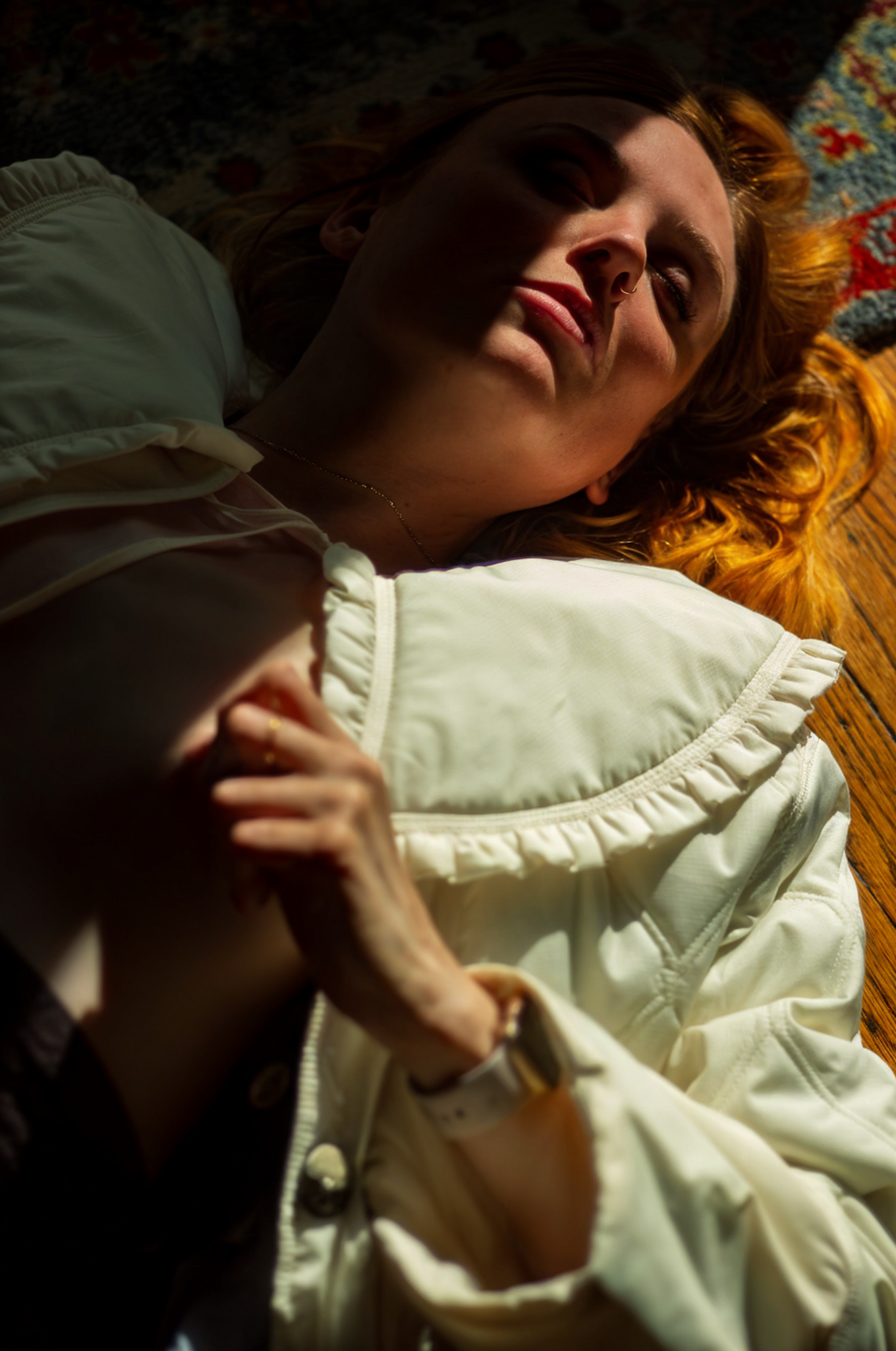 A woman with red hair lying on a colorful rug, with shadows cast across her face, wearing a light-colored jacket.