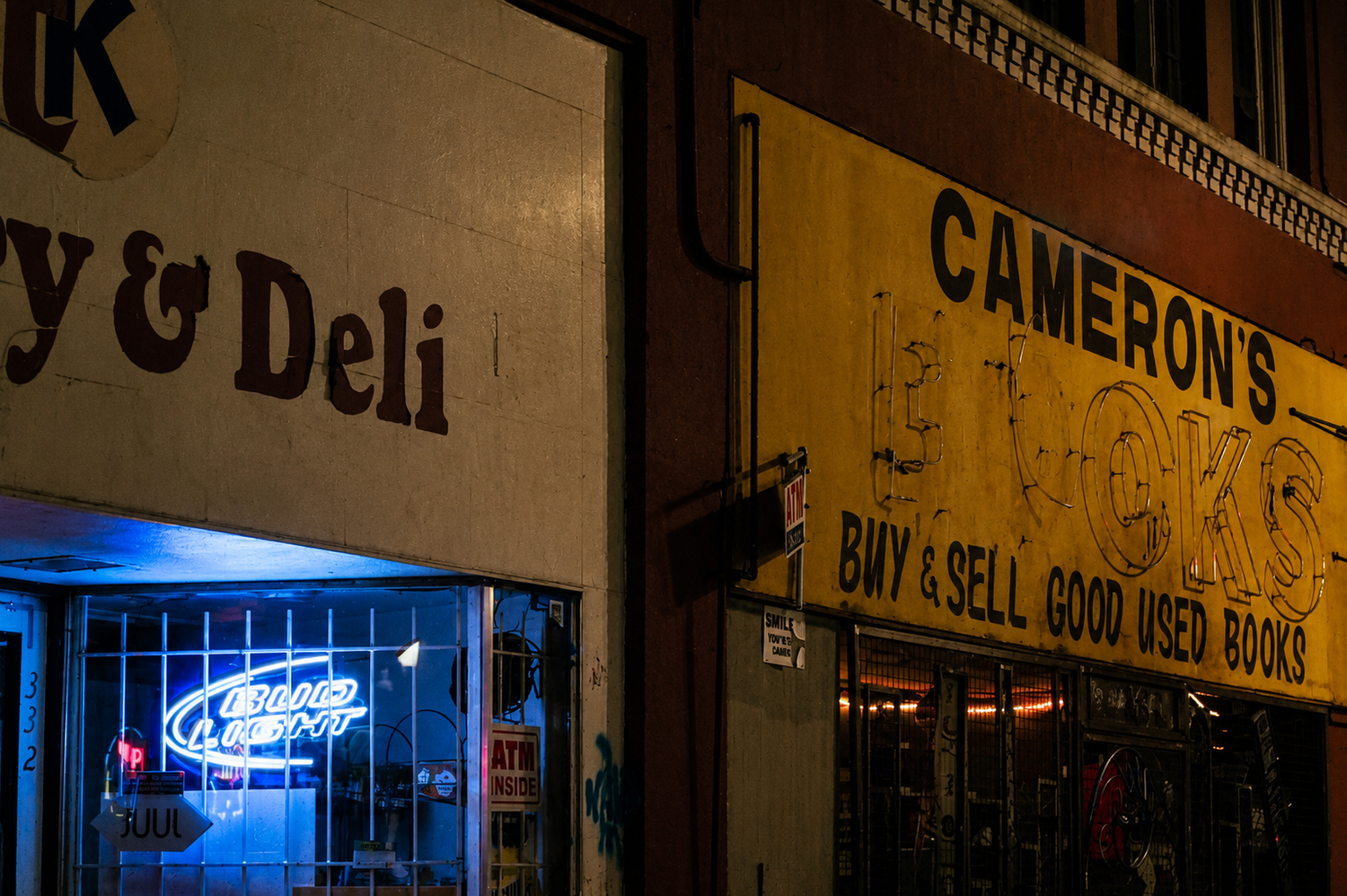 Nighttime street view showing a store with a sign that reads 'Cameron's' and 'Buy & Sell Good Used Books'. There is a neon sign inside the store that says 'Blood Light'.